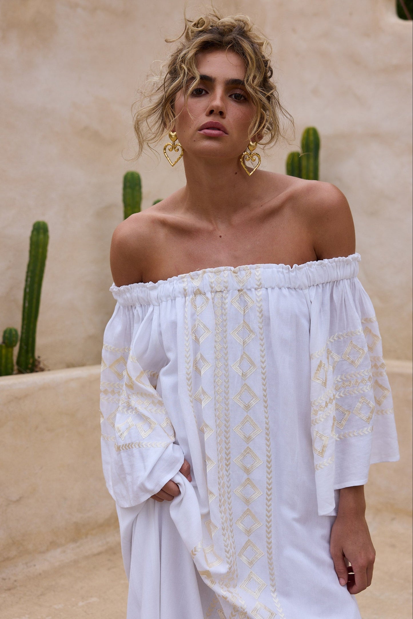 Woman wearing a white off-shoulder dress with gold patterns, gold heart shaped earrings in a desert setting.
