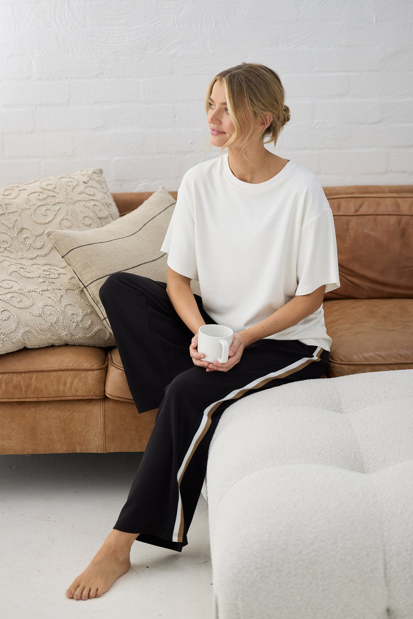 Woman sitting on a couch holding a mug, wearing a white shirt and black pants, with a neutral-colored wall in the background.
