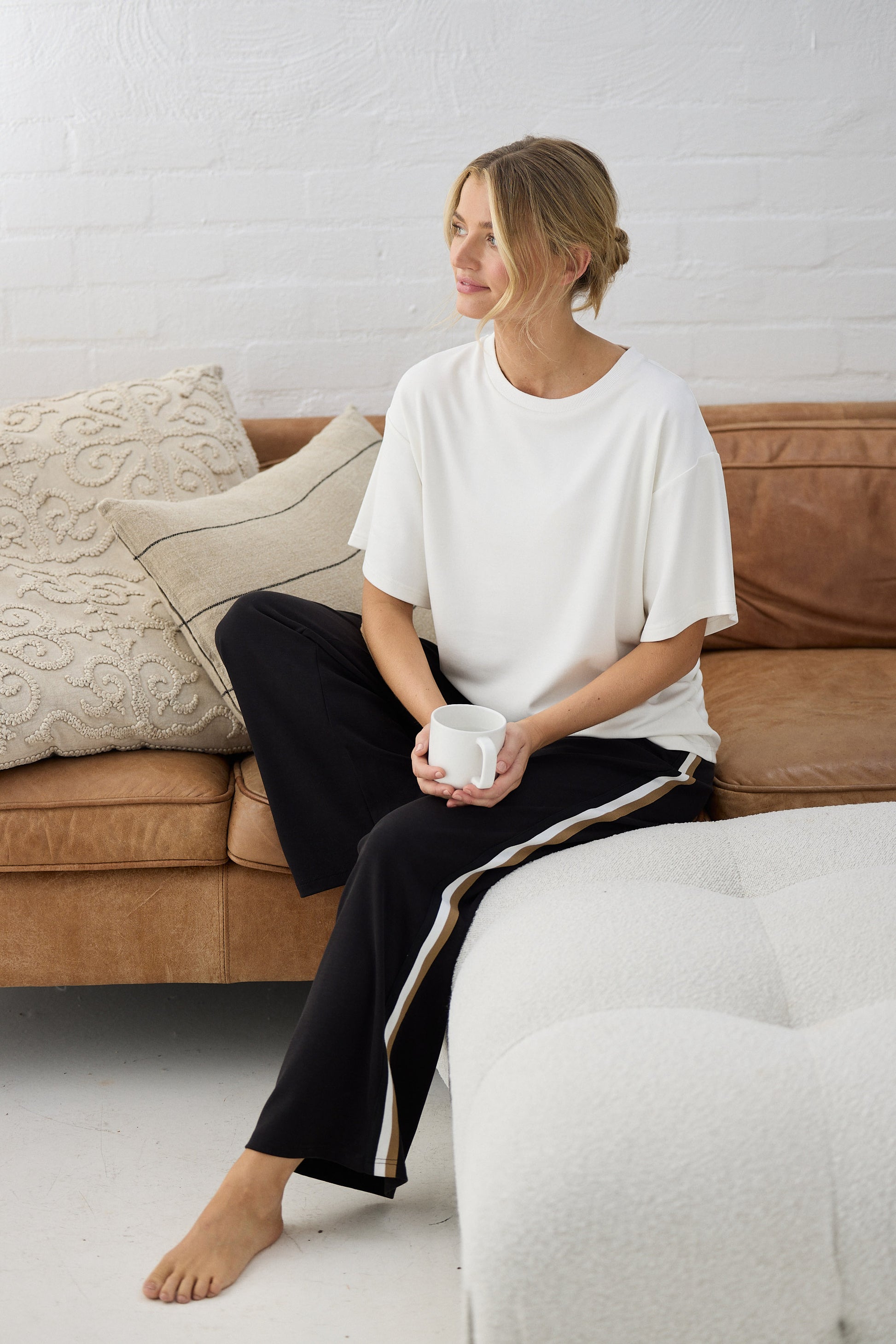 Woman sitting on a couch holding a mug, wearing a white shirt and black pants, with a neutral-colored wall in the background.