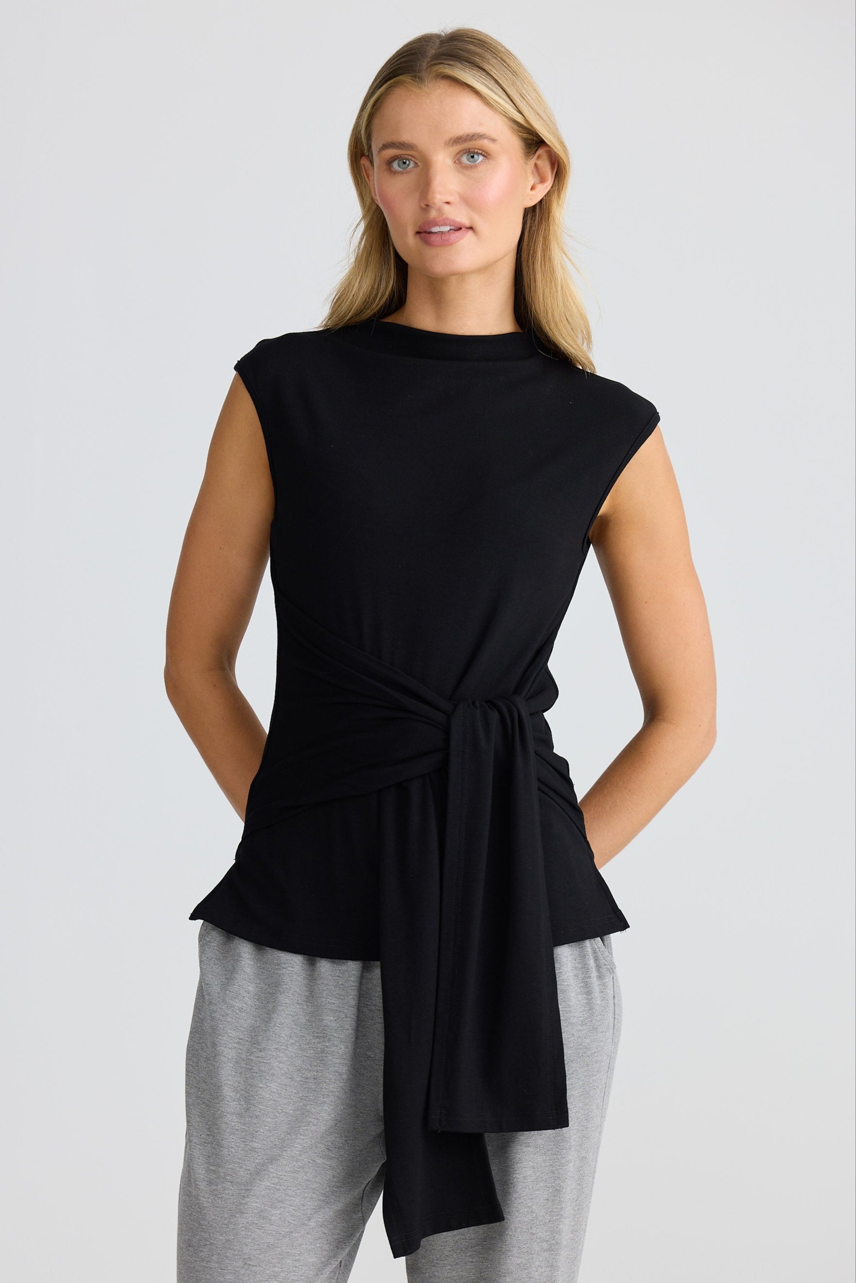 Woman wearing a black sleeveless top with a knot detail and gray pants on a white background