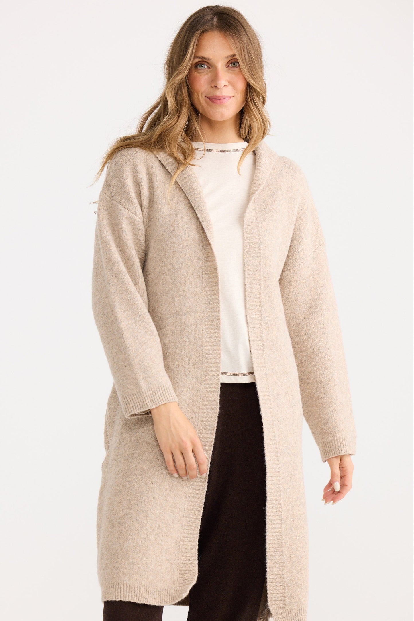 Woman wearing a beige cardigan over a white shirt and black pants on a white background