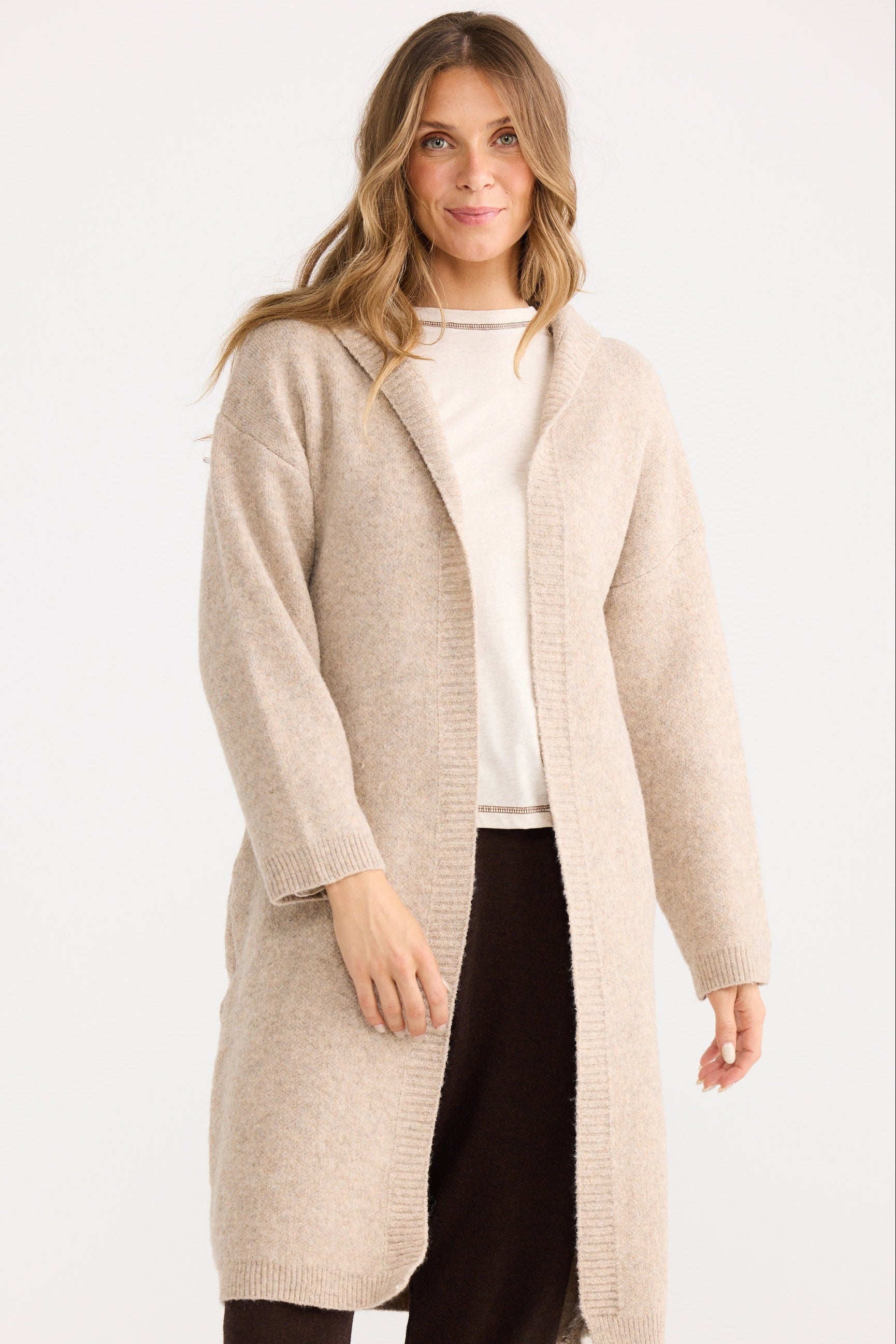 Woman wearing a beige cardigan over a white shirt and black pants on a white background