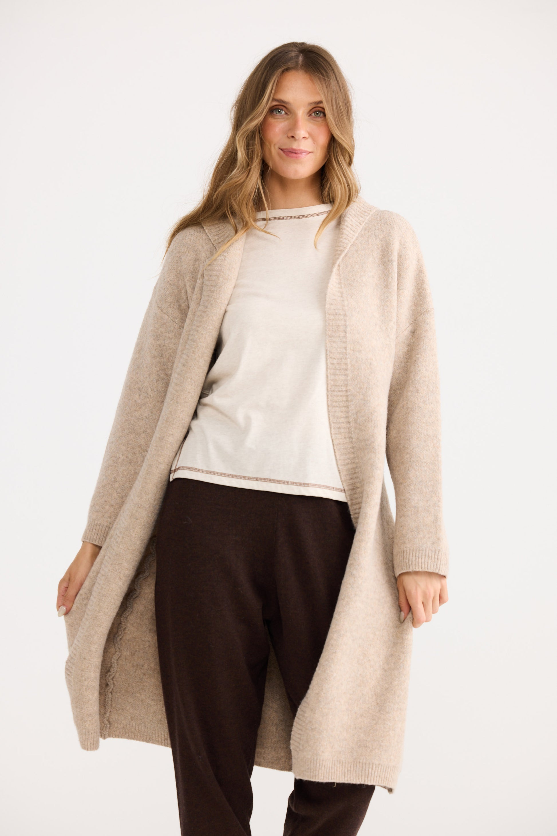 Woman wearing a long beige cardigan over a white top and dark pants on a white background
