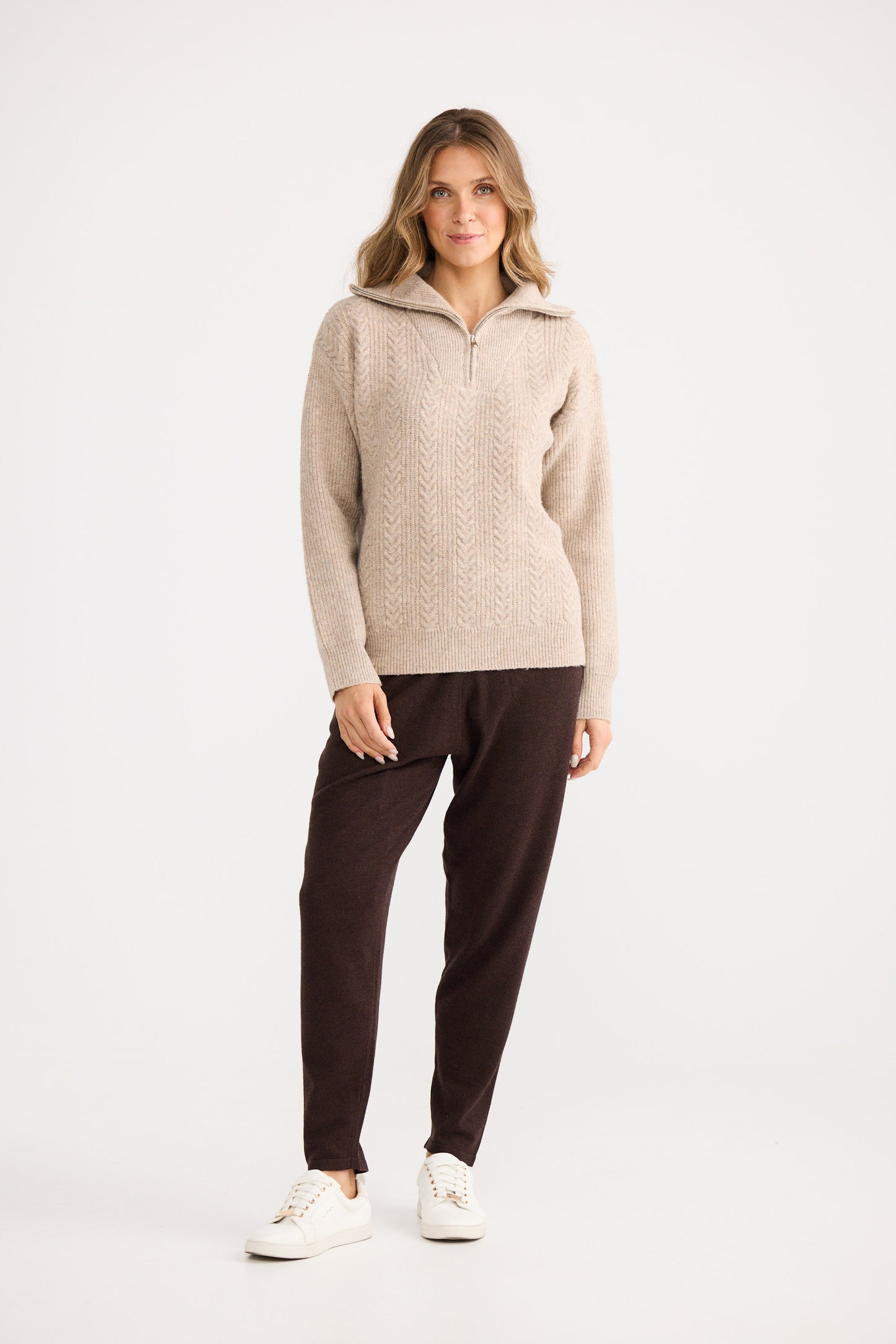 Woman wearing a beige sweater and dark pants on a white background