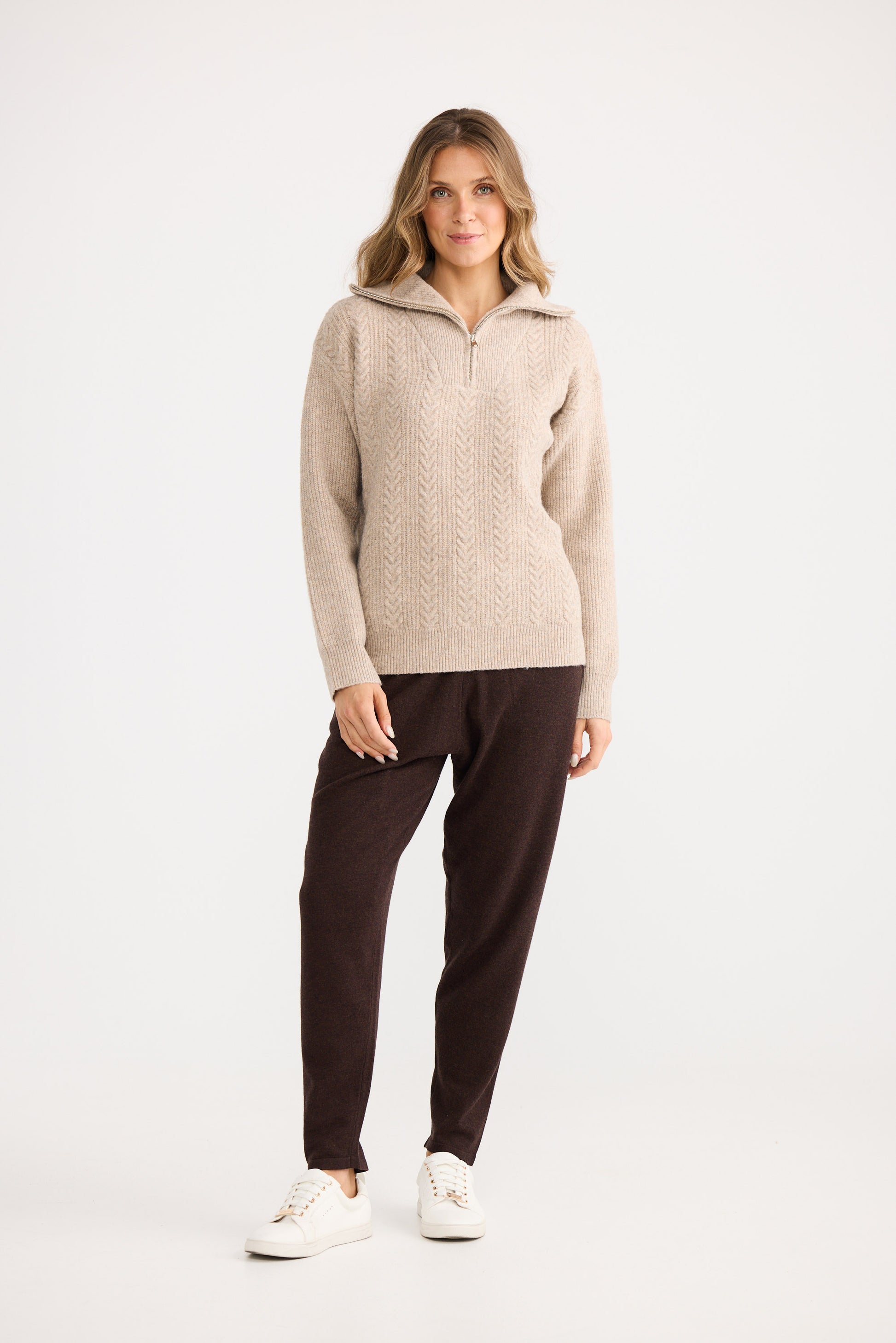 Woman wearing a beige sweater and dark pants on a white background