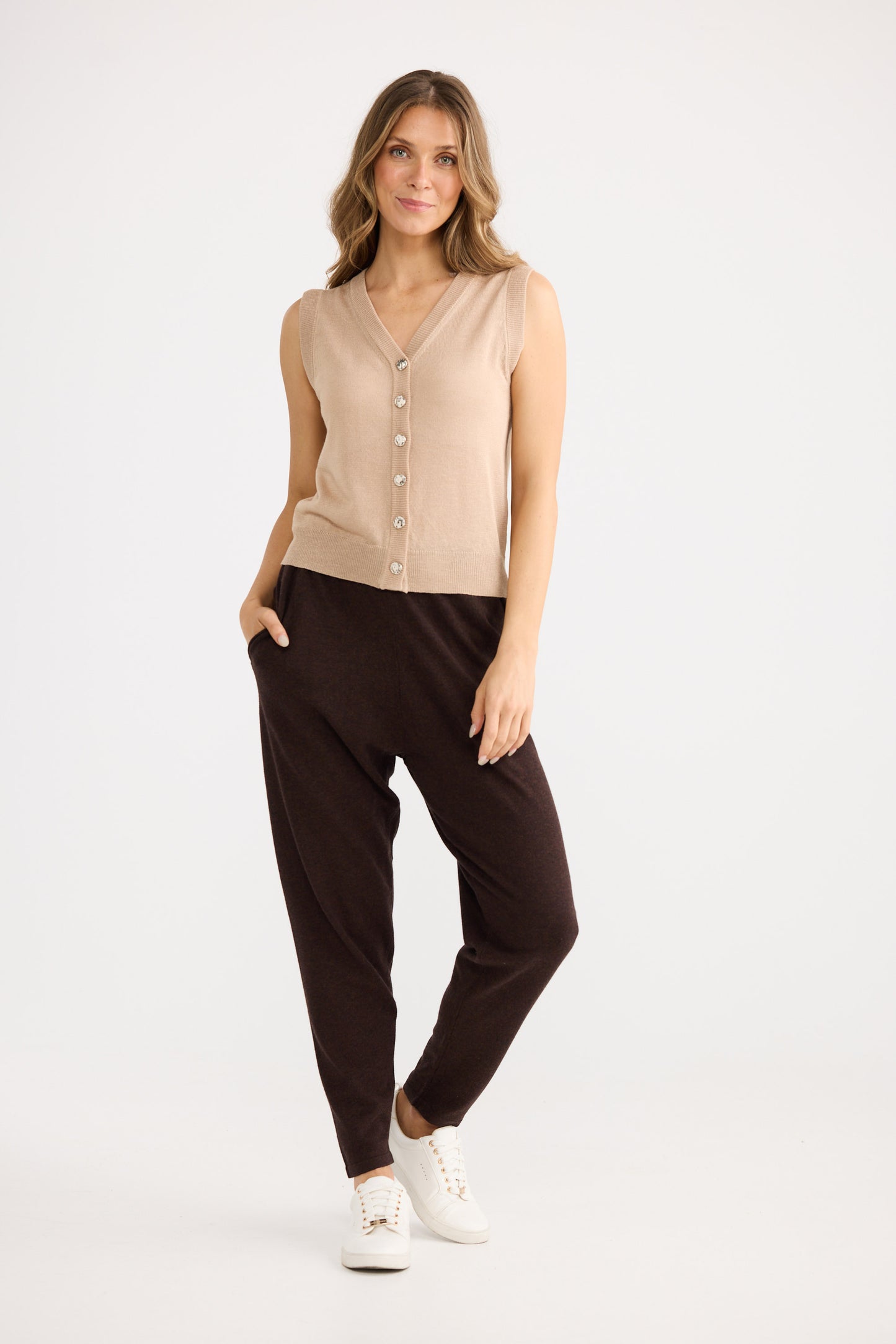 Woman wearing a beige sleeveless cardigan and brown pants on a white background