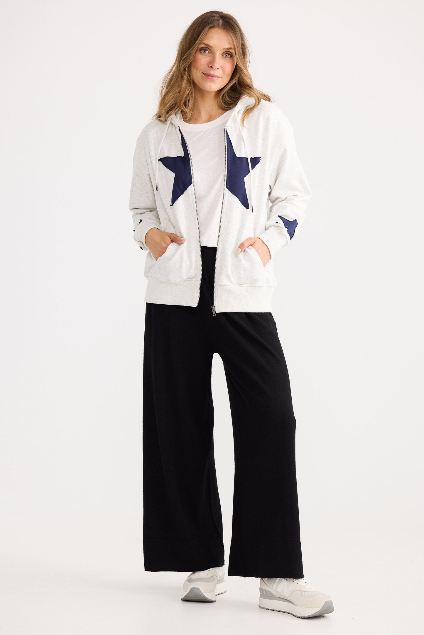 Woman wearing a white cardigan with star patterns, black pants, and white shoes on a white background