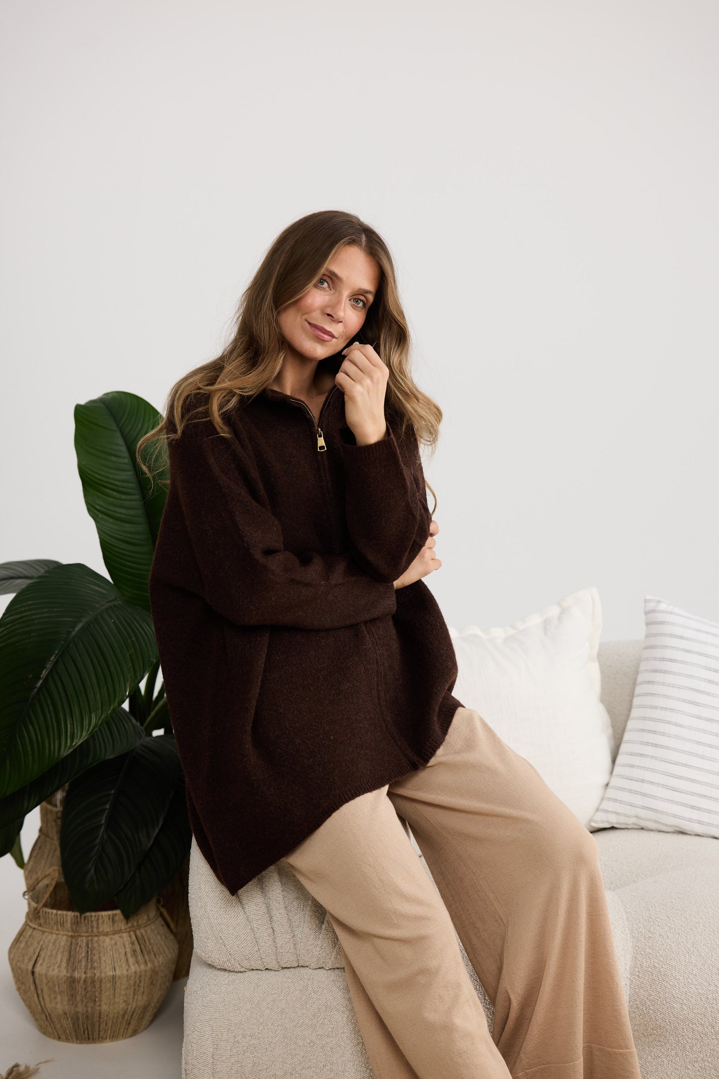 Woman sitting on a couch wearing a brown sweater and beige pants, with a white wall and plant in the background.