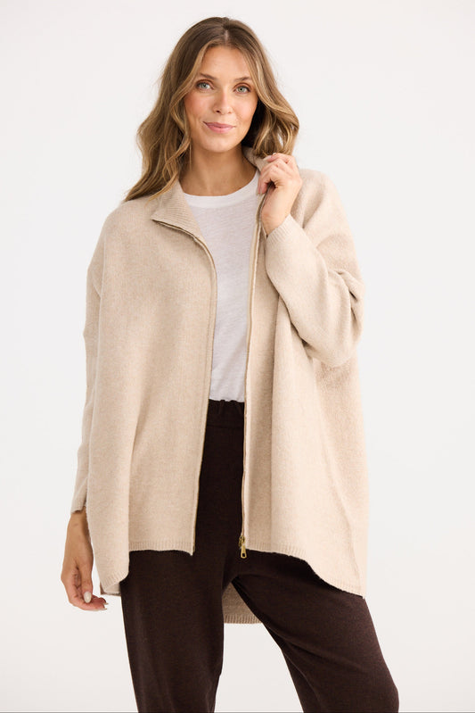 Woman wearing a beige coat over a white shirt and dark pants on a white background